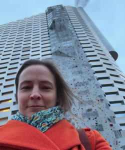 Me, a brunette woman, in an orange coat standing in front of a very tall building with smoke rising from the top.